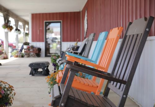 Colorful rocking chairs line a porch, with flowers and a red barn-style building in the background.