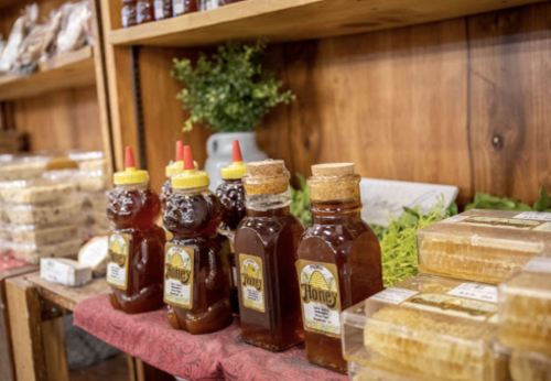 Bottles of honey in bear shapes on a wooden shelf, surrounded by honeycomb and greenery.