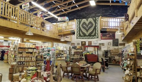 A cozy, rustic store interior with wooden beams, shelves filled with goods, and a large heart-patterned quilt hanging above.