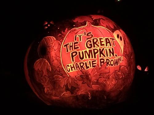 A carved pumpkin glowing in the dark, featuring the text "It's the Great Pumpkin, Charlie Brown" surrounded by leaves.
