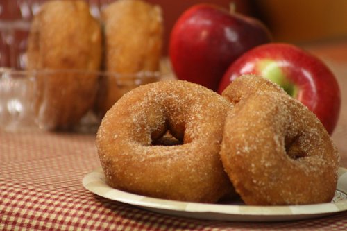 Two cinnamon sugar-coated donuts on a plate, with red apples and more donuts in the background.