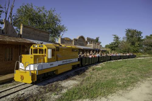 A yellow train with passengers on board, parked near a western-style building under a clear blue sky.