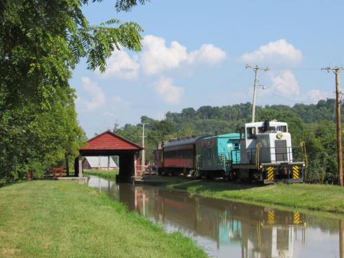 A train parked near a covered bridge, alongside a calm waterway, surrounded by greenery and blue skies.