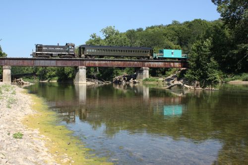 A train crosses a bridge over a calm river, surrounded by lush greenery and clear blue skies.