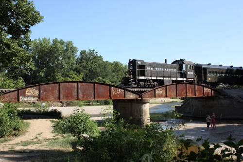 A vintage train crosses a rusted bridge over a river, surrounded by trees and greenery. Two people stand nearby.