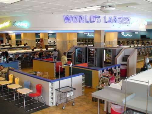 A spacious laundromat with colorful seating areas, laundry machines, and customers doing laundry. "World's Largest" sign visible.