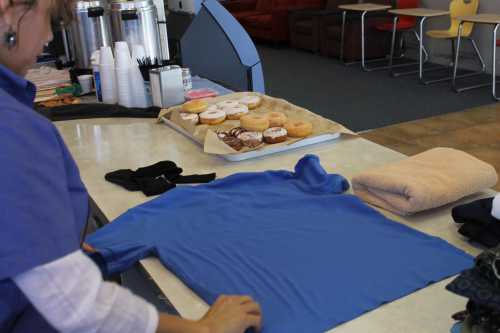 A person folds a blue shirt on a table next to a tray of donuts and various items like towels and utensils.