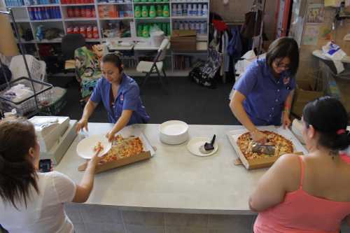 Two women serve pizza from boxes at a counter while a third woman takes a photo. The setting appears casual and communal.