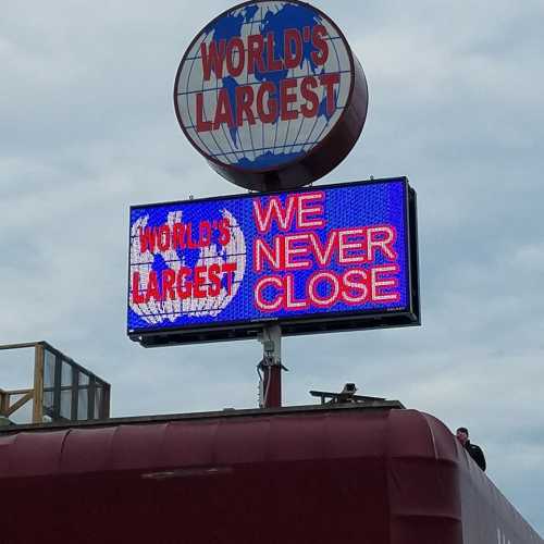 Sign reading "WORLD'S LARGEST" and "WE NEVER CLOSE" with a globe graphic, set against a cloudy sky.