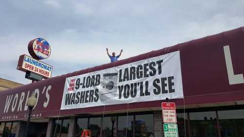 A person stands on a laundromat roof, celebrating a banner that advertises new 9-load washers as the largest available.