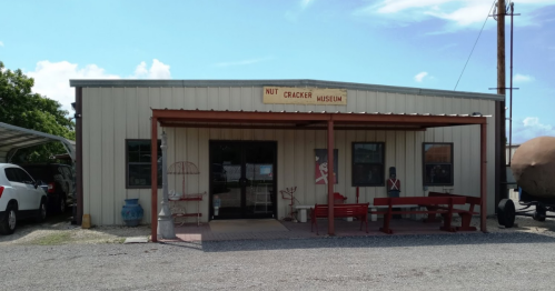 Exterior of a metal building labeled "Nut Cracker Museum," featuring a porch and outdoor seating. Clear blue sky above.