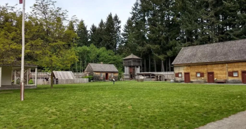 A grassy area with wooden buildings and a lookout tower, surrounded by tall trees under a cloudy sky.