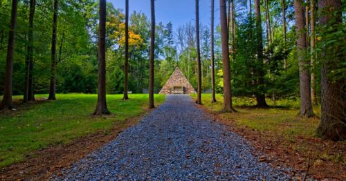 A gravel path leads through tall trees to a pyramid-shaped structure in a serene forest setting.