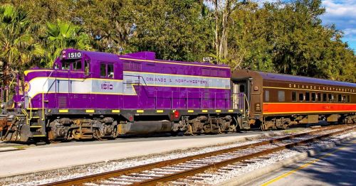 A purple and white locomotive with a vintage passenger car parked beside the tracks under a clear blue sky.