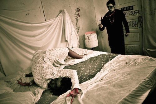 A person in a nightgown lies on a bed, while another figure in dark clothing approaches with a cross.