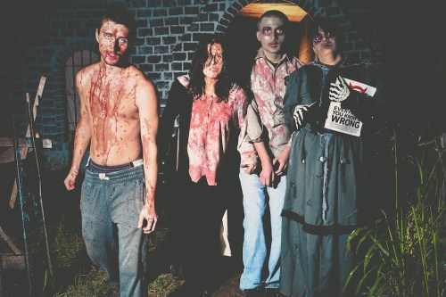 Four people in horror-themed costumes, covered in fake blood, standing in a dimly lit, eerie outdoor setting.