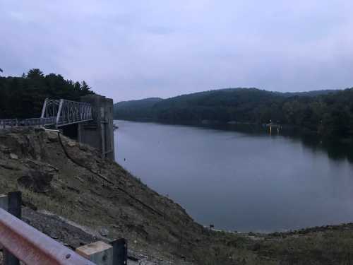 A calm river flows beside a dam, surrounded by wooded hills under a cloudy sky at dusk.