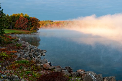 A serene lakeside scene with colorful autumn trees, mist rising from the water, and rocky shoreline.