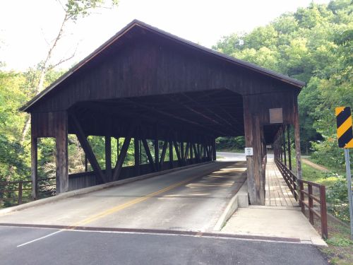 A wooden covered bridge spans a road, surrounded by lush green trees and a clear sky.