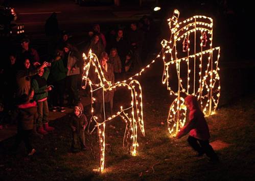 A festive scene with a glowing horse and carriage made of lights, surrounded by a crowd enjoying the display.