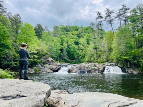 A person stands on a rock by a river, surrounded by lush green trees and a waterfall in the background.