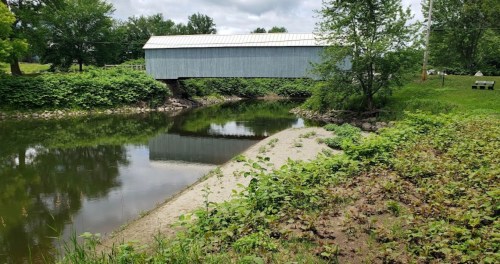 A covered bridge spans a calm river, surrounded by lush greenery and a sandy bank under a cloudy sky.