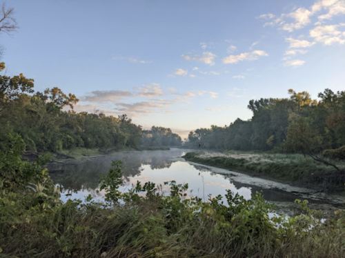A serene river scene at dawn, surrounded by lush greenery and mist rising from the water under a clear sky.