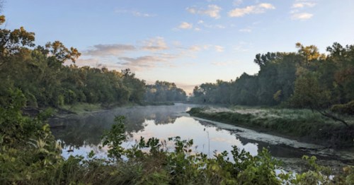 A serene river scene at dawn, surrounded by lush greenery and mist rising from the water.