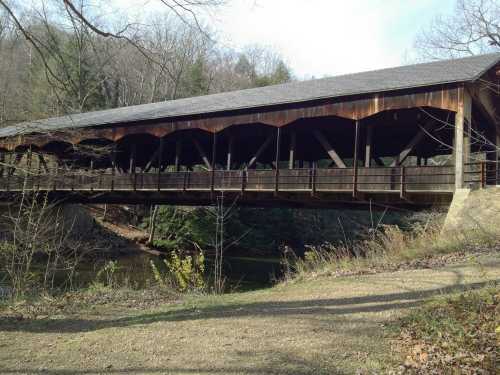 A wooden covered bridge spans a river, surrounded by trees and a grassy area under a clear sky.