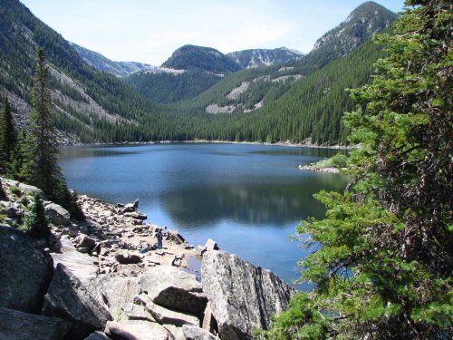 A serene mountain lake surrounded by lush green trees and rocky shores, with mountains in the background under a clear sky.