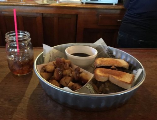 A metal tray with toast, potatoes, and a small bowl of sauce, alongside a mason jar filled with iced tea.