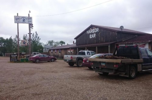 A rustic bar named "Harbor Bar" with parked vehicles and a gravel lot, surrounded by trees under a cloudy sky.