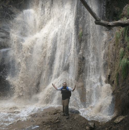 A person stands in front of a large waterfall, surrounded by rocky terrain and splashing water.