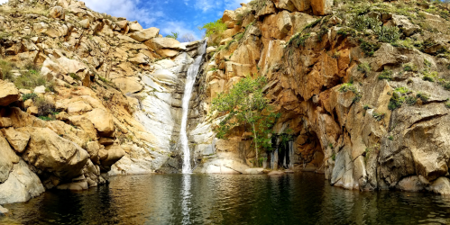 A serene waterfall cascades into a tranquil pool, surrounded by rocky cliffs and lush greenery under a blue sky.