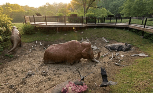 A prehistoric scene with life-sized dinosaur and mammoth sculptures among bones, set in a grassy area with a wooden deck.