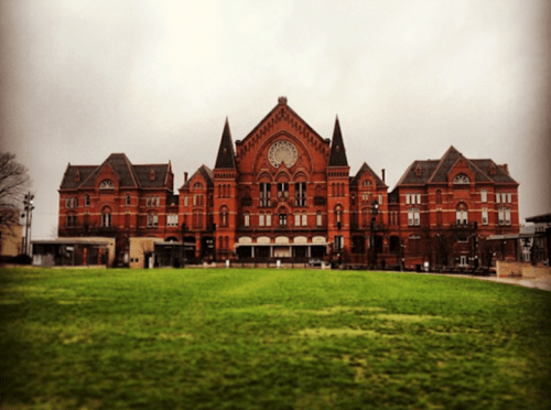 Historic red brick building with intricate architecture, set against a cloudy sky and green lawn in the foreground.