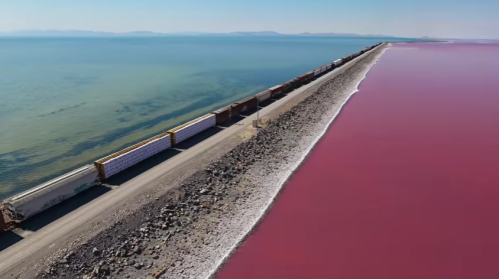 Aerial view of a train track along a shoreline with vibrant pink water and rocky beach.