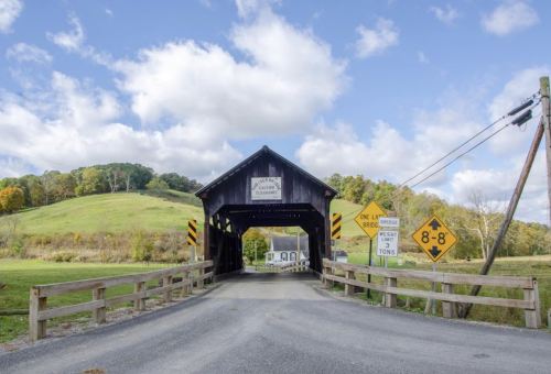A covered bridge with a wooden structure, surrounded by green hills and a cloudy sky, with road signs nearby.