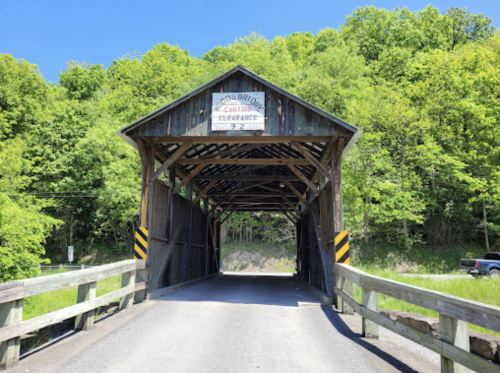 A wooden covered bridge with a sign, surrounded by lush green trees and a clear blue sky.
