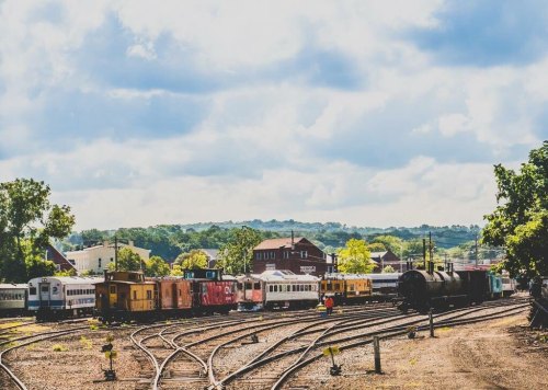 A train yard with colorful train cars and tracks under a cloudy sky, surrounded by greenery and buildings in the background.