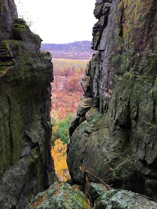 A narrow rock crevice with moss-covered walls, revealing a view of a valley with autumn foliage in the distance.