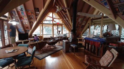 Cozy wooden cabin interior with sloped ceilings, a living area, and large windows overlooking greenery.