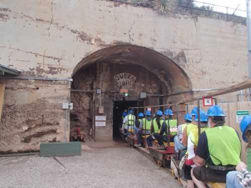 A group of people in hard hats waits to enter a tunnel marked "Queen 1915," with mining equipment visible.