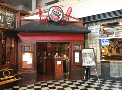 A cozy restaurant entrance with a red roof, featuring a sign and a woman at the host stand, surrounded by checkered flooring.