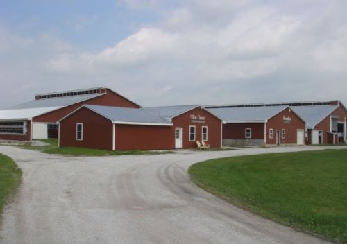 A red barn complex with multiple buildings, surrounded by a gravel path and green grass under a cloudy sky.
