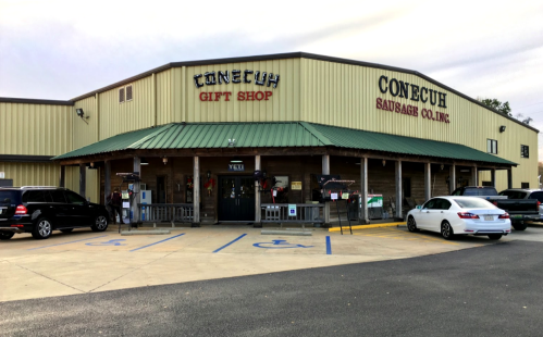 Exterior of Conecuh Sausage Co. gift shop, featuring a green roof and parking area with several cars.