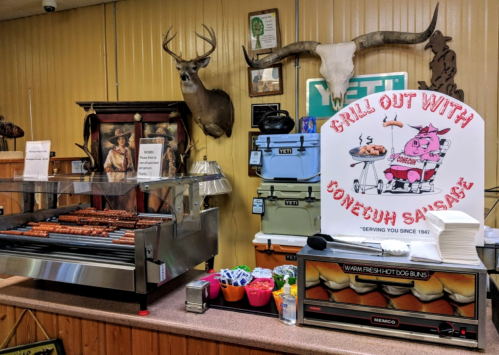 A rustic food stand featuring a grill with sausages, a sign promoting "Bonecuh Sasae," and mounted animal heads on the wall.