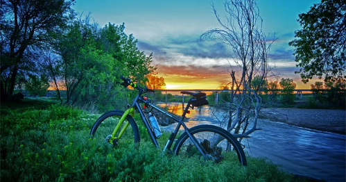 A bike rests by a riverbank at sunset, surrounded by lush greenery and colorful skies.
