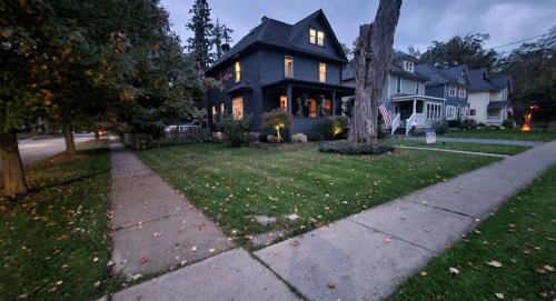 A dark blue house with a porch, surrounded by trees and a well-kept lawn, at dusk with streetlights glowing.