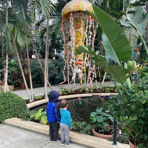 Two children stand by a pond in a greenhouse, gazing at a colorful jellyfish decoration hanging above.